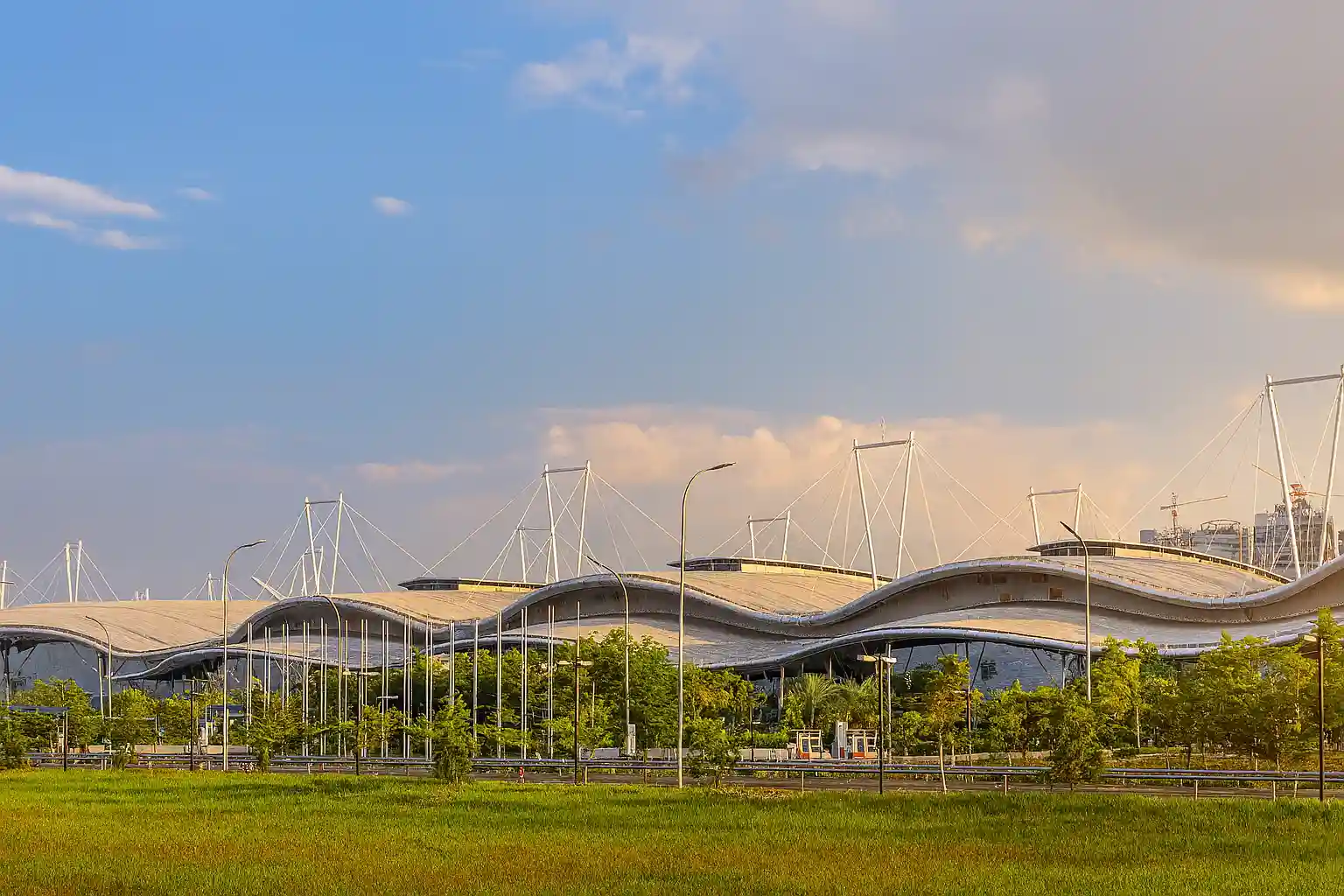 Tampak gedung ICE BSD dengan atap bergelombang menjelang sore hari, dikelilingi pepohonan hijau dan langit cerah keemasan sebagai lokasi Event Otomotif Gaikindo 2025.